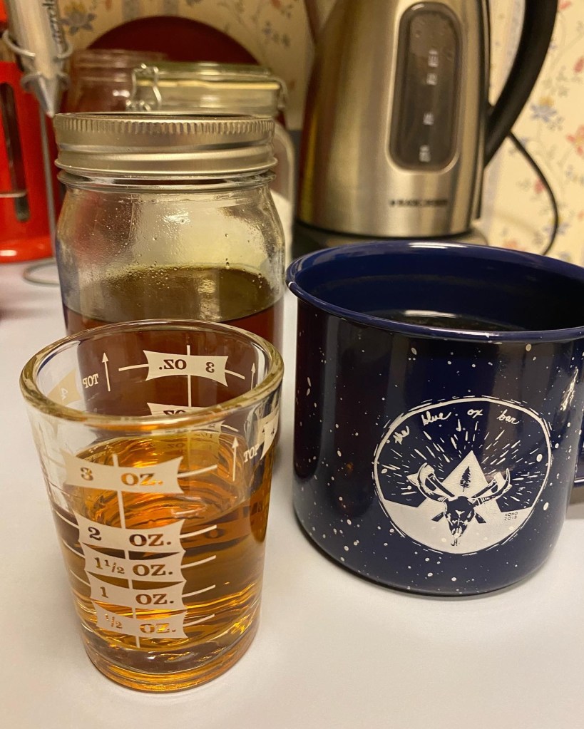 The blue metal white-speckled mug sits next to a measuring shot glass with amber liquid (sotol) and a mason jar of honey