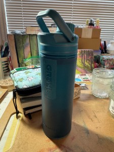 A blue water bottle with a spout and handle, with the words Grayl down the side and Ultrapress at the bottom. It is translucent plastic with a rubberized cap and ring around the bottom for traction.